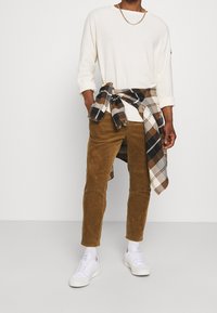 Man in een wit overhemd met lange mouwen, bruine korduroy broek, witte sneakers en een geruite shirt om zijn taille geknoopt, tegen een effen achtergrond.