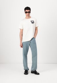 Tricou alb cu mânecă scurtă, având un grafic închis pe piept, purtat cu blugi drepti de culoare albastră deschisă și pantofi negri.