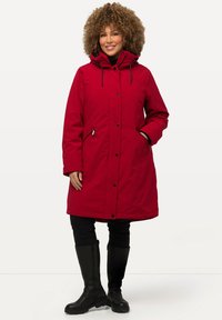Red, insulated parka with adjustable hood, front snap closure, and two side pockets. Model wearing black pants and knee-high boots.