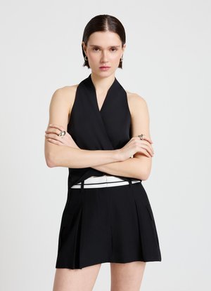 Femme aux cheveux courts et foncés portant un top noir sans manches à col halter et une jupe noire avec ceinture blanche, debout les bras croisés devant un fond gris.