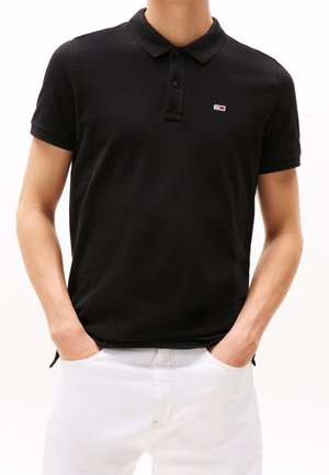 Uomo che indossa una polo nera a maniche corte con un piccolo logo sul petto e pantaloni bianchi, in piedi con le mani in tasca su uno sfondo bianco.