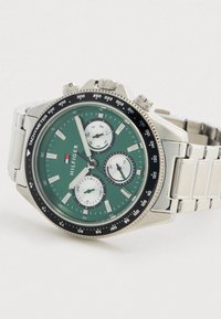 Ezüst színű fém karkötőóra zöld számlappal, három segédszámlappal, tachiméter gyűrűvel és láncos fém szíjjal, fehér háttéren szögben elhelyezve.