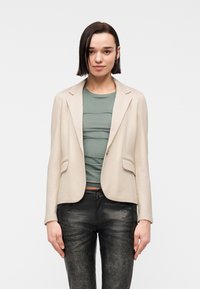 Femme aux cheveux courts et foncés, portant un blazer beige, un haut vert et un pantalon noir texturé, debout devant un fond blanc uni.