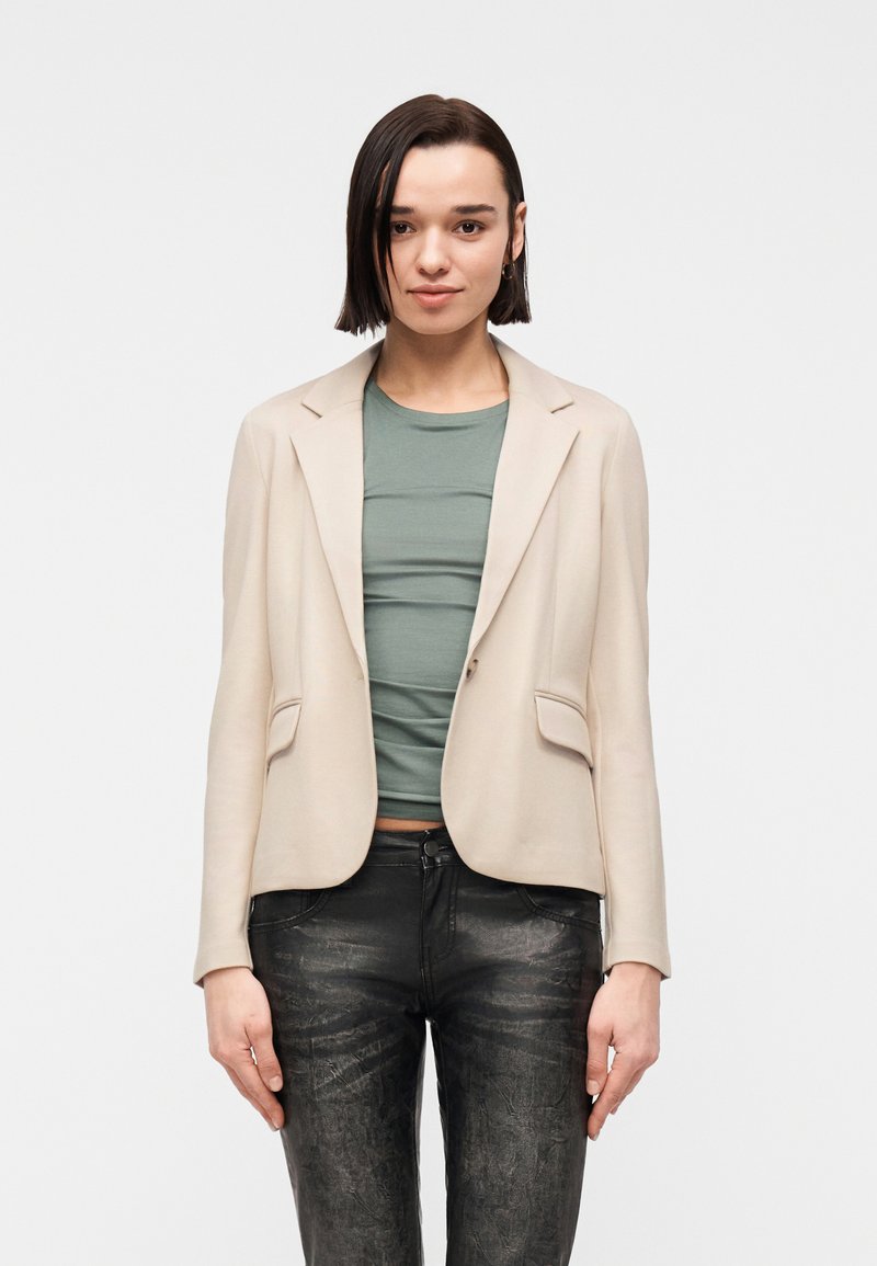 Femme aux cheveux courts et foncés, portant un blazer beige, un haut vert et un pantalon noir texturé, debout devant un fond blanc uni.