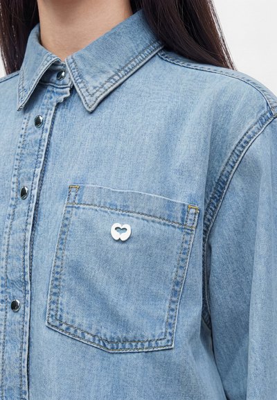 Gros plan d'une personne portant une chemise en jean bleu clair avec des boutons-pression argentés et une poche avant ornée d'une petite épingle en forme de cœur argentée.