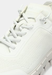 Λευκό αθλητικό παπούτσι με υφή από πλεκτό τοπίο στο επάνω μέρος και λείες λεπτομέρειες. Διαθέτει ανοιχτόχρωκα κορδόνια και μοτίβο στη σόλα. Το λογότυπο είναι ορατό στην γλώσσα του παπουτσιού.