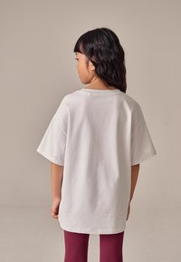 Weißes kurzärmliges T-Shirt mit lockerer Passform, aus Baumwolle, kombiniert mit gerippten kastanienbraunen Leggings. Glatte Textur und Rückansicht angezeigt.