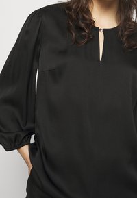 Blouse noire en tissu lisse, dotée d'un décolleté en trou de serrure, de manches bouffantes et d'un léger éclat, avec une petite poche sur le côté.