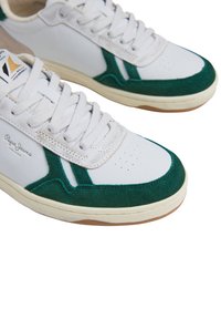 Bela teniska z zelenimi semiš detajli in gumijastimi podplati. Ima teksturirane panele, okroglo konico in ravne vezalke. Vidna blagovna znamka.