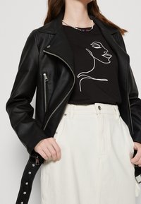 Czarna skórzana kurtka motocyklowa z srebrnymi suwakami, noszona na czarnej koszulce z białym rysunkiem. W połączeniu z białymi spodniami o wysokim stanie.