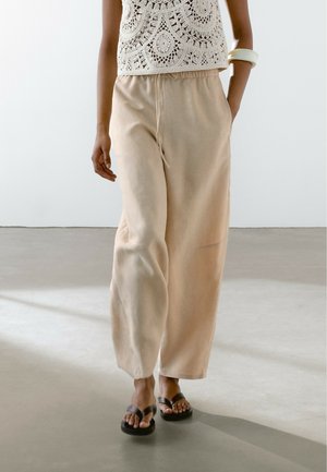 Femme portant un pantalon beige à jambes larges, un top sans manches en crochet, des sandales noires et un bracelet blanc, debout sur un sol en béton lisse.