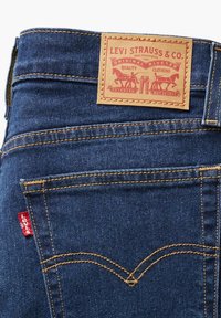 Jeans de mezclilla azul con un parche de cuero marrón claro que presenta una marca en rojo y dorado, costuras amarillas y una pequeña etiqueta roja en el bolsillo trasero.