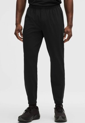 Mand iført sorte joggerbukser med elastisk talje og sorte sportssko, stående mod en ensartet lys baggrund.