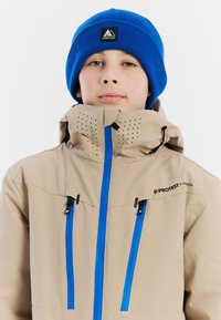 Beige sneeuwjack met blauwe ritsen en accenten, voorzien van perforaties in de kraag, gedragen met een blauwe gebreide beanie.
