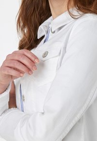 Žena s dlouhými vlasy, oděná v bílém sakovém kabátu s kapsami, drží jednu ruku poblíž oblasti hrudníku.