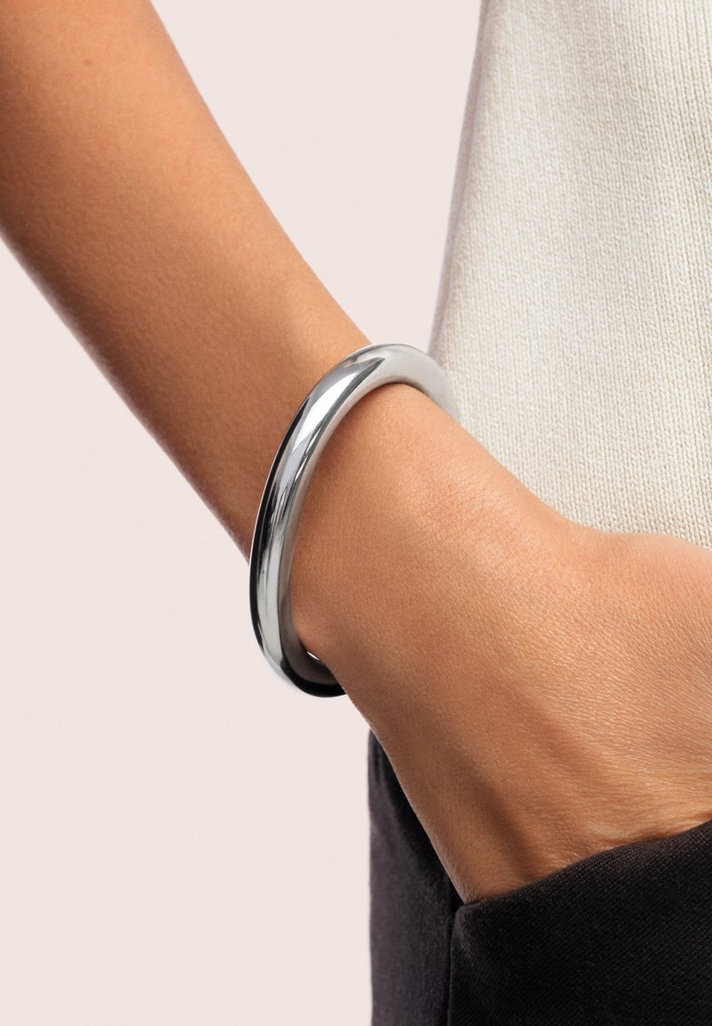 Brazalete de metal plateado reposando sobre una mano, con una superficie lisa y brillante y forma redondeada. La muñeca es parcialmente visible.