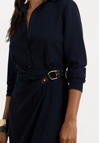 Jūras zila pogu kleita ar garām piedurknēm, piesprausta ar melnu jostu ar zelta aksesuāriem. Gluda auduma drapējuma dizains.