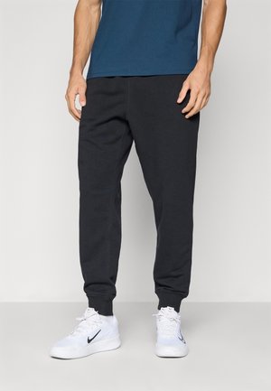 Man wearing black jogger pants, white Nike sneakers, and a blue shirt standing against a plain background.
