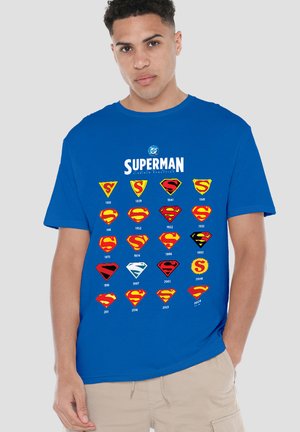 Junger Mann trägt ein blaues T-Shirt, das die Entwicklung der Superman-Logos von 1938 bis 2024 in Reihen auf der Vorderseite zeigt.