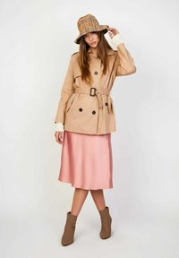 Trench beige con cinturón y botones negros, combinado con una falda de satén rosa y un sombrero tipo bucket de cuadros marrón. Botines marrones completan el conjunto.