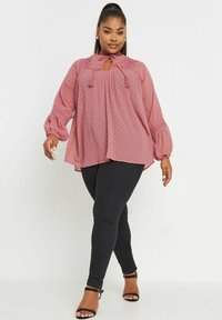 Blusa a maniche lunghe color rosa pallido con design a pois, scollatura a occhiello, colletto arricciato e polsini elasticizzati, abbinata a jeans skinny neri e tacchi.