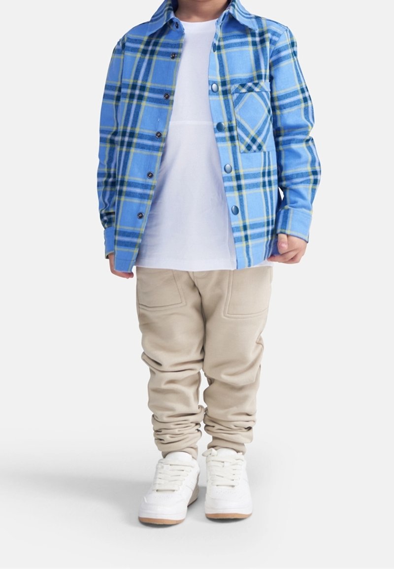 Chemise à carreaux bleue avec des accents verts, t-shirt blanc, joggings beige, et baskets blanches avec semelles en gomme. La tenue comprend une poche avant sur la chemise.
