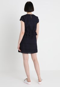 Nainen, jolla on lyhyet tummat hiukset, pukeutuneena laivansinisessä reikäkankaasta valmistetussa mekossa, jossa on lyhyet hihat, ja valkoisissa platform-sandaaleissa, seisoo valkoista taustaa vasten.