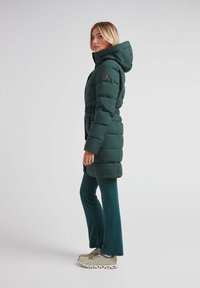 Abrigo acolchado verde con capucha, diseño de acolchado, cinturón ajustable en la cintura, combinado con pantalones acampanados verdes y zapatillas ligeras.