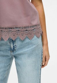 Top morado con borde de encaje, que presenta intrincados patrones florales; combinado con jeans de lavado claro, mostrando un ajuste casual.