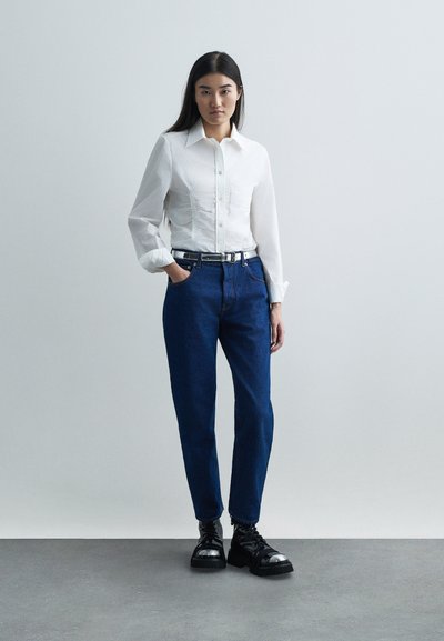 Moteris stovi prieš šviesią fono, apsirengusi balta marškinėlių su sagomis, tamsiai mėlynomis džinsomis su diržu ir juodais batais su metaliniais pirštais.