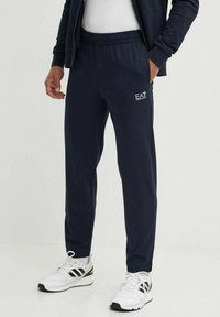 Pantaloni tuta blu navy con elastico in vita, tasche laterali e dettaglio del logo EA7. Realizzati in tessuto liscio, vestibilità affusolata e polsini in rete.
