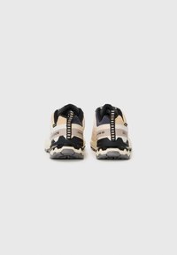 Chaussures de trail running avec une tige beige, des accents noirs et une semelle texturée. Présente un tirant distinct à l'arrière et des détails de logo.