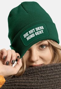 Green knit beanie with a textured design and a rectangular patch featuring white text that reads "DIE MIT DEM HUND GEHT." Textured brown scarf visible.