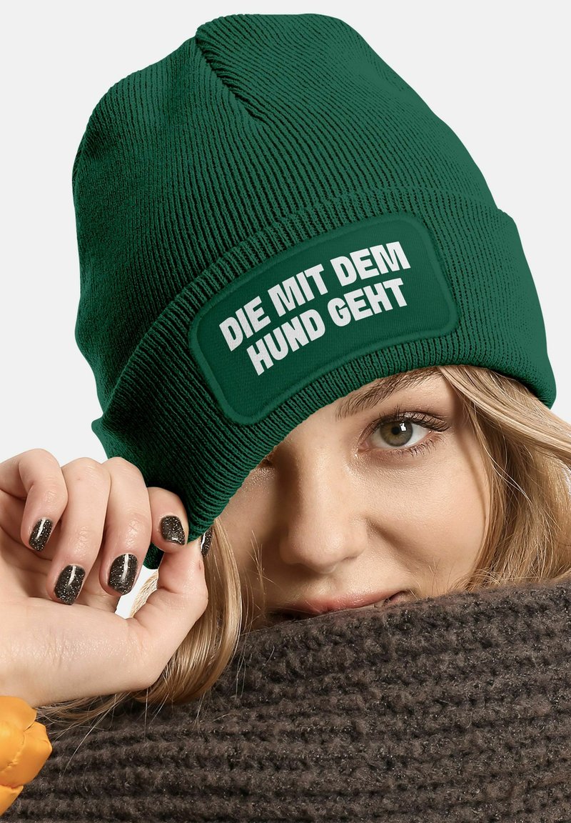 Green knit beanie with a textured design and a rectangular patch featuring white text that reads "DIE MIT DEM HUND GEHT." Textured brown scarf visible.
