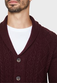 Burgundský pletený kardigan s šálovým límcem, s kabelovým vzorem, dvěma knoflíky a bílým tričkem s kulatým výstřihem pod ním.