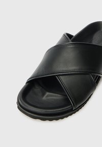 Svart skinnslippersandal med krysset stroppdesign, teksturert fotseng og fleksibel gummisåle. Glatt overflate med minimal søm.