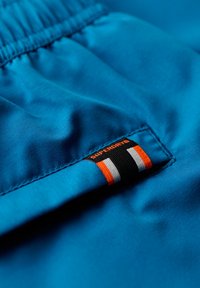 Blauwe stof met een gladde textuur, voorzien van een oranje en zwarte label met de tekst "SUPERDRY" en drie verticale strepen in oranje, zwart en wit.