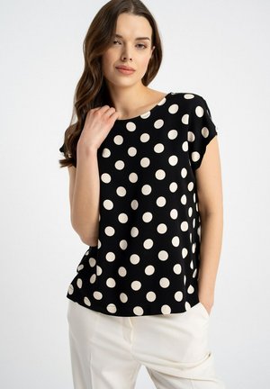 Kobieta z długimi brązowymi włosami, ubrana w czarną bluzkę na krótki rękaw z dużymi białymi grochami i białe spodnie, stoi na tle jednolitego jasnego tła.