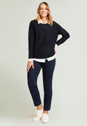 Marineblauer Strickpullover mit strukturiertem Muster und kleinen Verzierungen, getragen über einem weißen Hemd mit Kragen, kombiniert mit dunklen Jeans und weißen Turnschuhen.