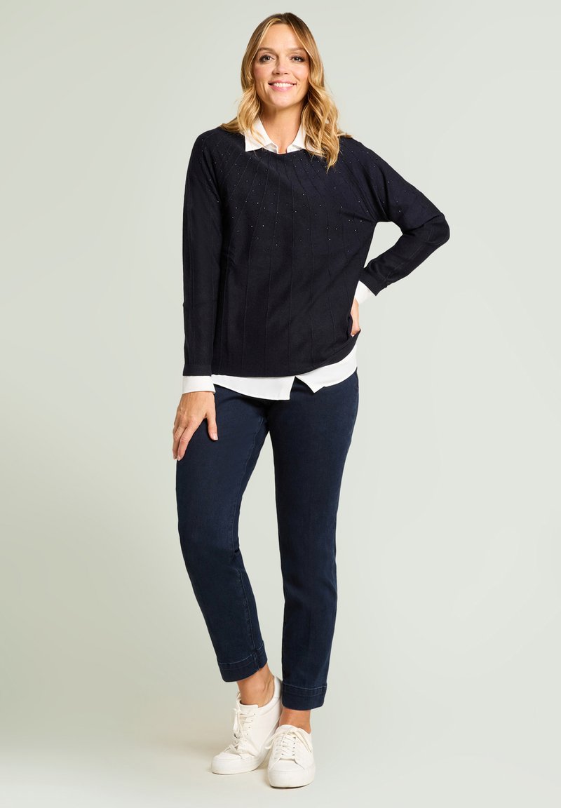 Granatowy dzianinowy sweter z teksturowanym wzorem i małymi ozdobami, nałożony na białą koszulę z kołnierzykiem, zestawiony z ciemnymi dżinsami i białymi sneakersami.