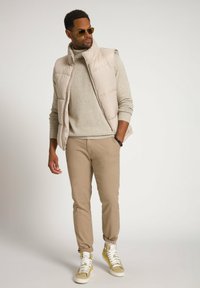 Gilet imbottito beige sopra un maglione beige chiaro, abbinato a pantaloni beige e sneakers bianche con accenti gialli. Visibilità chiara delle texture e dei colori.