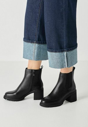 Schwarze Lederstiefeletten mit Blockabsatz, getragen zu dunkelblauen, umgekrempelten Jeans, die hellblauen Innenstoff zeigen.