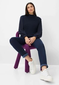 Marineblauwe geribde trui met ballonmouwen, donkerblauwe opgerolde broek en witte sneakers, zittend op een paarse stoel tegen een witte achtergrond.