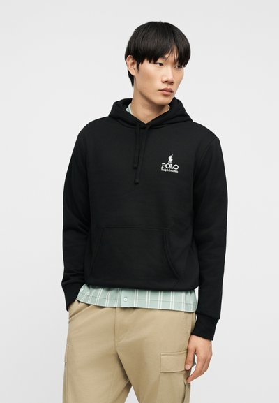 Νέος άνδρας που φοράει μαύρο φούτερ Polo Ralph Lauren πάνω από ανοιχτό μπλε πουκάμισο με καρό και μπεζ παντελόνι cargo, στέκεται μπροστά σε απλό φόντο.