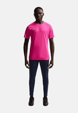 Junger Mann, der nach vorne schaut, trägt ein knallpinkes Sportshirt mit kurzen Ärmeln, dunkle Sporthose und schwarze Sportschuhe.