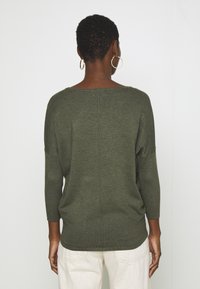 Grön stickad tröja med vid halsringning, trekvartsärmar och ribbad textur, med en central sömdekor på baksidan.