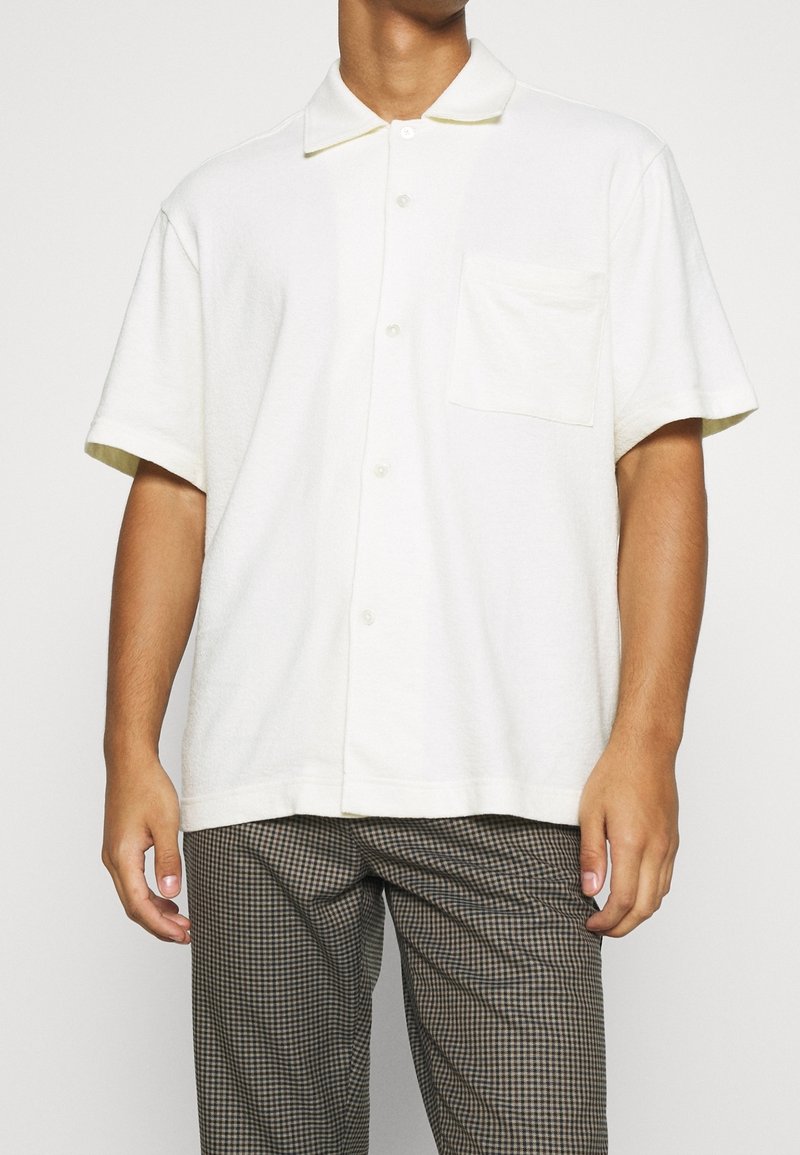 Koszula z krótkim rękawem i kołnierzykiem w odcieniu ecru, wykonana z materiału o teksturowym wykończeniu, z kieszenią na piersi i zapięciem na guziki, noszona z wzorzystymi spodniami.