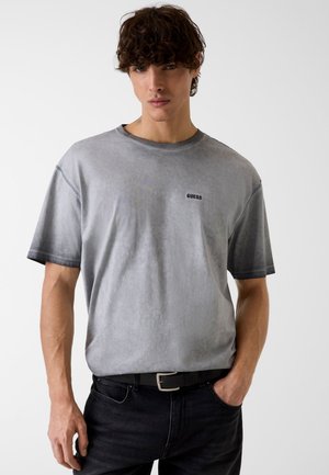 Jonge man met krullend donker haar, gekleed in een vergrijsd grijs Guess T-shirt en zwarte jeans, staand met één hand in zijn broekzak tegen een witte achtergrond.