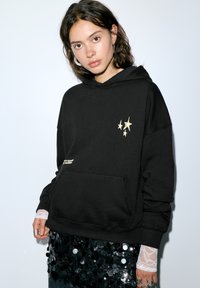 Zwarte hoodie met een losse pasvorm, voorzien van gouden sterrenprint en een voorzak. Draagt over een zwarte paillettenrok met kanten manchetten.