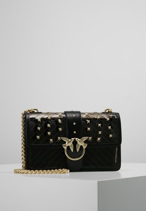 Musta tikattu nahkalaukku, jossa on kullanväriset mehiläisnappulat, kullanvärinen lintuklipsi ja paksu kullanvärinen ketjuhihna, valkoisella jalustalla vaaleanharmaata taustaa vasten.